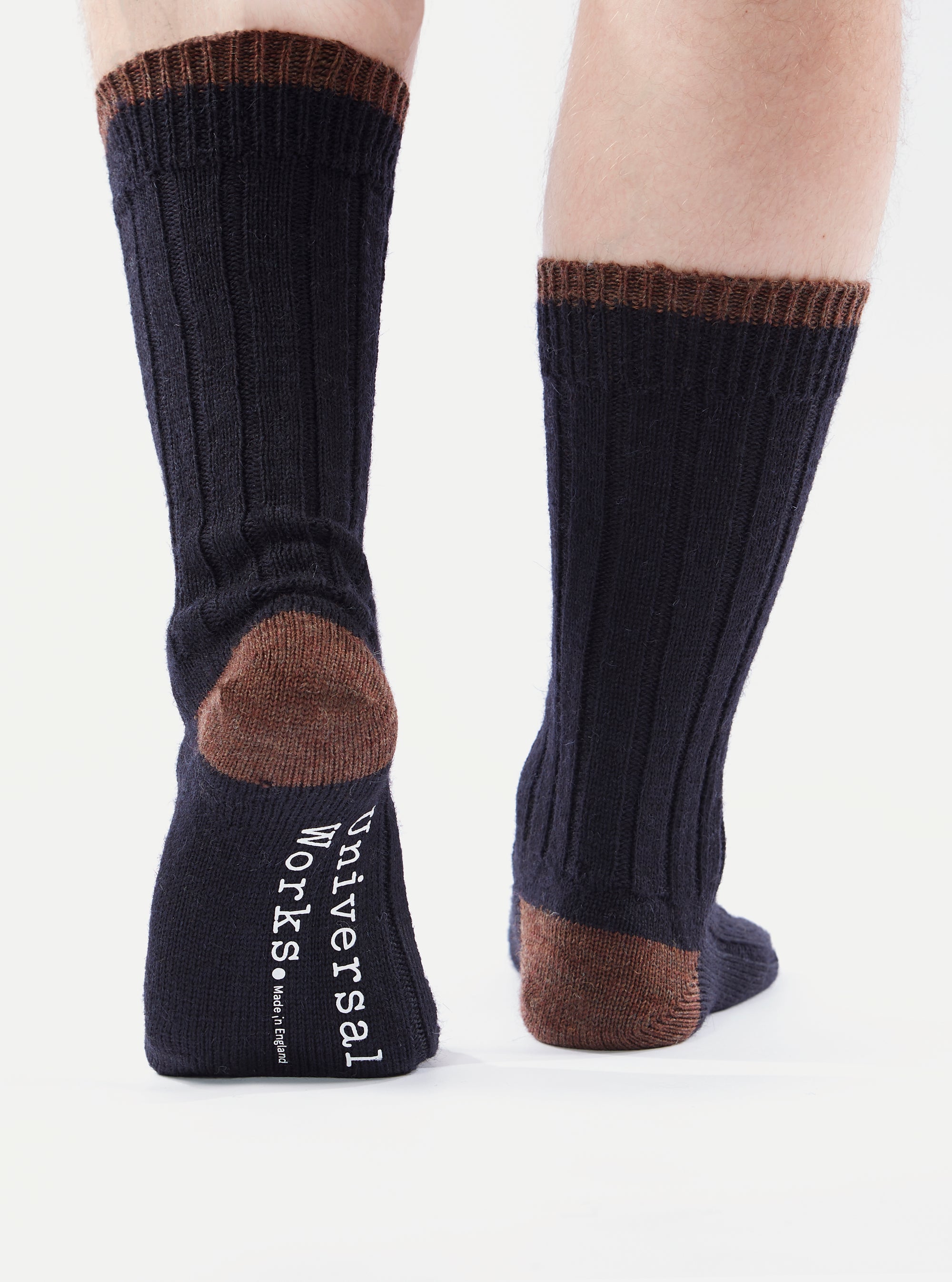 Close-up of a person wearing Navy Wool Hike Sock. The socks are navy with brown accents on the heel and cuff. The background is a bright white.