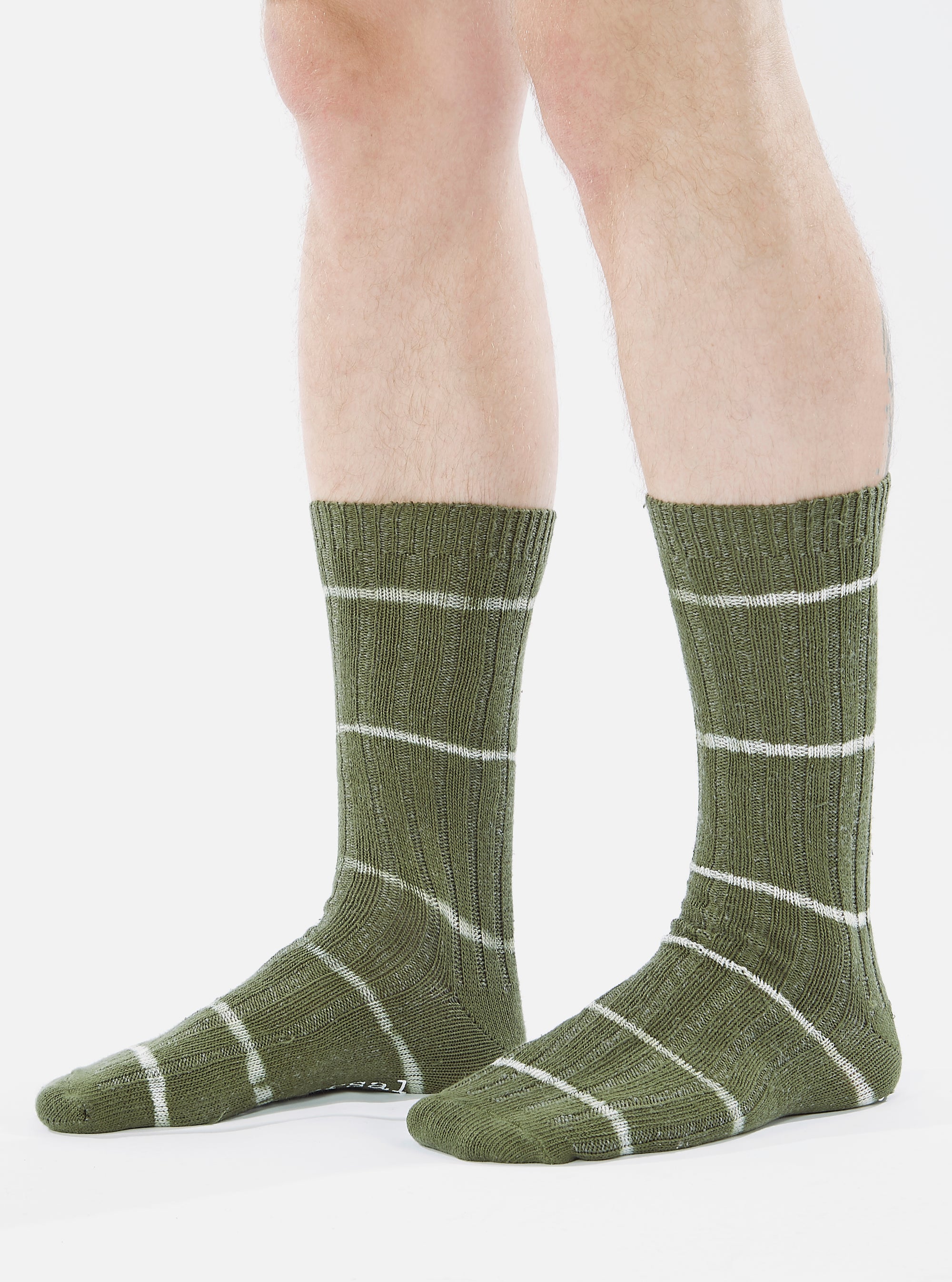 Close-up of a model wearing Olive Tie Dye Knit Slub Sock. The olive green socks have white horizontal stripes. Shot against a white background.