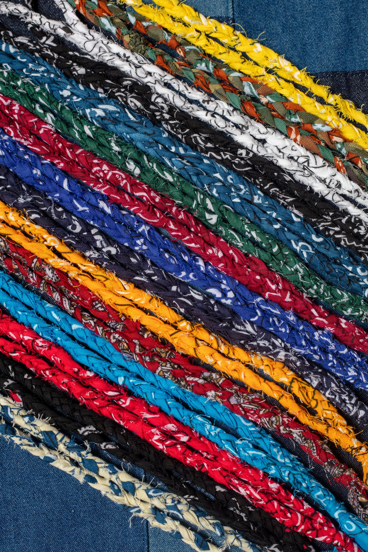 Close-up of the Hanami of Rome Bandana Necklace/Bracelet #5. Braided strips of colorful bandana fabric (red, blue, yellow, black, white) are arranged diagonally on a denim background.
